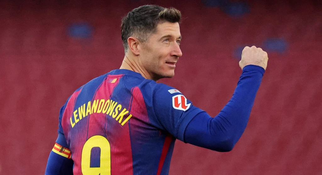Piłkarz w koszulce FC Barcelony z nazwiskiem Lewandowski i numerem 9 na plecach, wykonujący gest triumfu na tle stadionu.