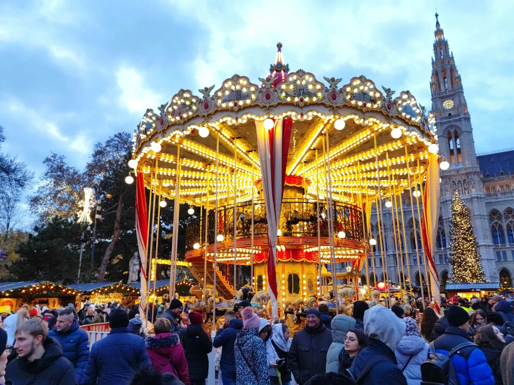 Na jarmarku w Wiedniu nie jest taniej niż na jarmarkach w Polsce Karuzela ozdobiona licznymi światełkami otoczona przez tłum ludzi. W tle widoczne wysokie, oświetlone drzewko świąteczne i fragment zabytkowego budynku z wieżą.