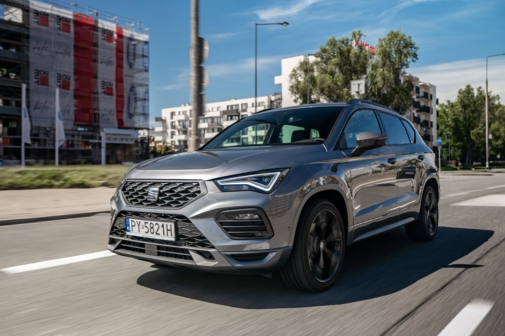 Nowoczesny, srebrny SUV marki Seat poruszający się po miejskiej drodze, w tle budynki mieszkalne i drzewa, wyraźne oznakowanie poziome jezdni, słoneczna pogoda i niebieskie niebo.