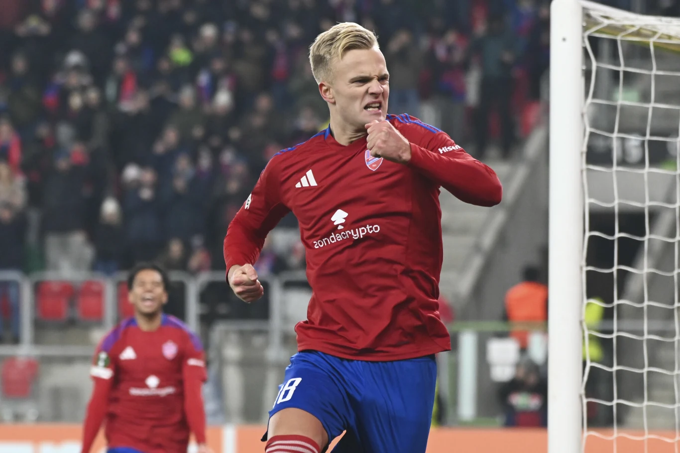 Piłkarz w czerwonym stroju drużyny świętuje zdobytego gola trzymając pięść przy klatce piersiowej, w tle widać innego zawodnika oraz kibiców na trybunach.