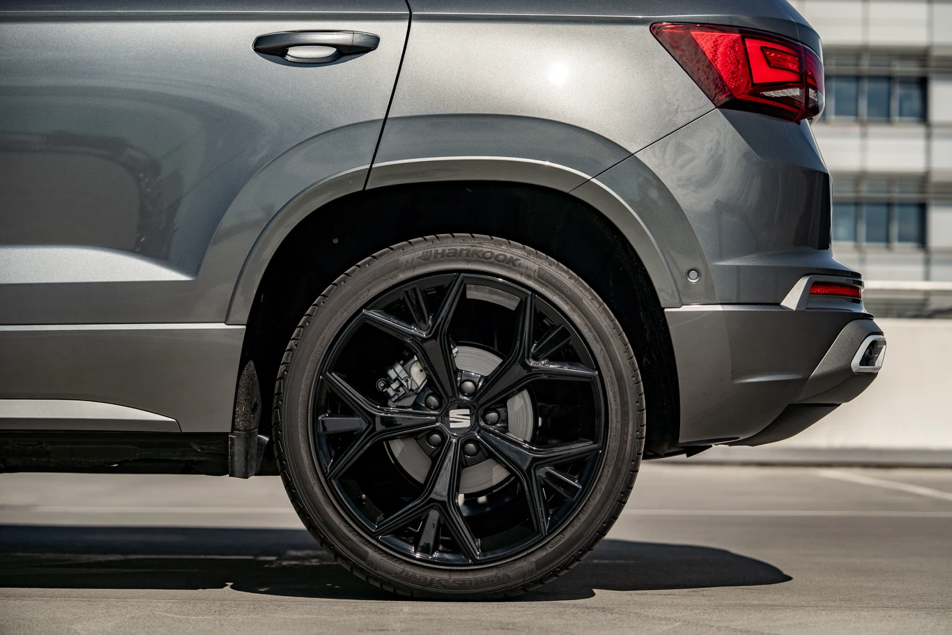 Szczegółowy widok tylnej części szarego samochodu osobowego stojącego na betonowej powierzchni, z wyeksponowanym czarnym felgą aluminiową oraz fragmentem lampy tylnej, karoserii i zderzaka.