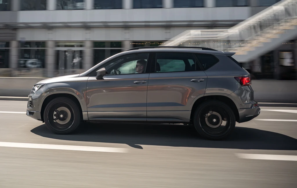 Srebrny SUV poruszający się po miejskiej drodze przy przeszklonym budynku i schodach w tle.