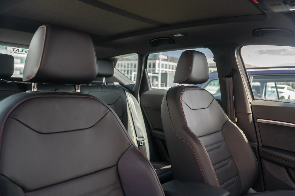 Wnętrze samochodu z widokiem na dwa przednie fotele pokryte czarną tapicerką z czerwonym obszyciem oraz lekko uchylone drzwi po stronie pasażera. Przez okno widoczne elementy innych pojazdów i budynki.