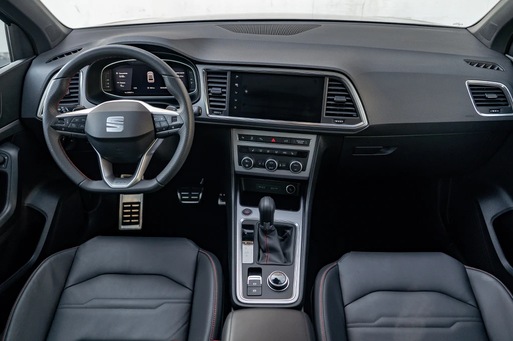 Kokpit nowoczesnego samochodu marki Seat, widoczna kierownica z logo, deska rozdzielcza z dużym ekranem, centralny panel z przyciskami i dźwignią zmiany biegów oraz dwa skórzane fotele przednie.