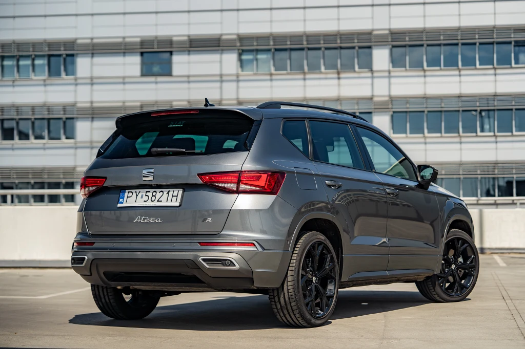 Srebrny SUV marki Seat Ateca na parkingu przy nowoczesnym budynku, widoczny tył pojazdu z czarnymi felgami i tablicą rejestracyjną.