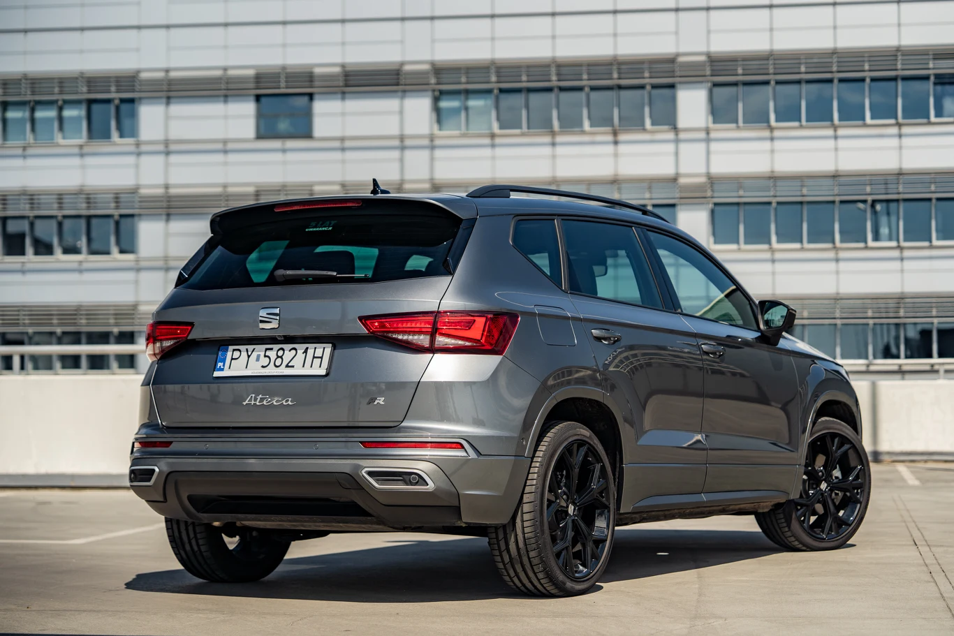 Srebrny SUV marki Seat Ateca na parkingu przy nowoczesnym budynku, widoczny tył pojazdu z czarnymi felgami i tablicą rejestracyjną.