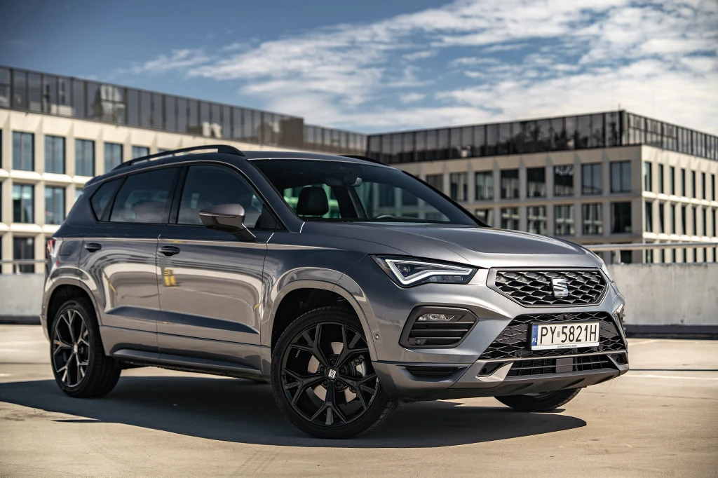 Nowoczesny, srebrny SUV marki Seat Ateca ustawiony na parkingu na tle wielopiętrowych, nowoczesnych budynków biurowych, wyraźne refleksy światła na karoserii, widoczne czarne felgi i tablica rejestracyjna.