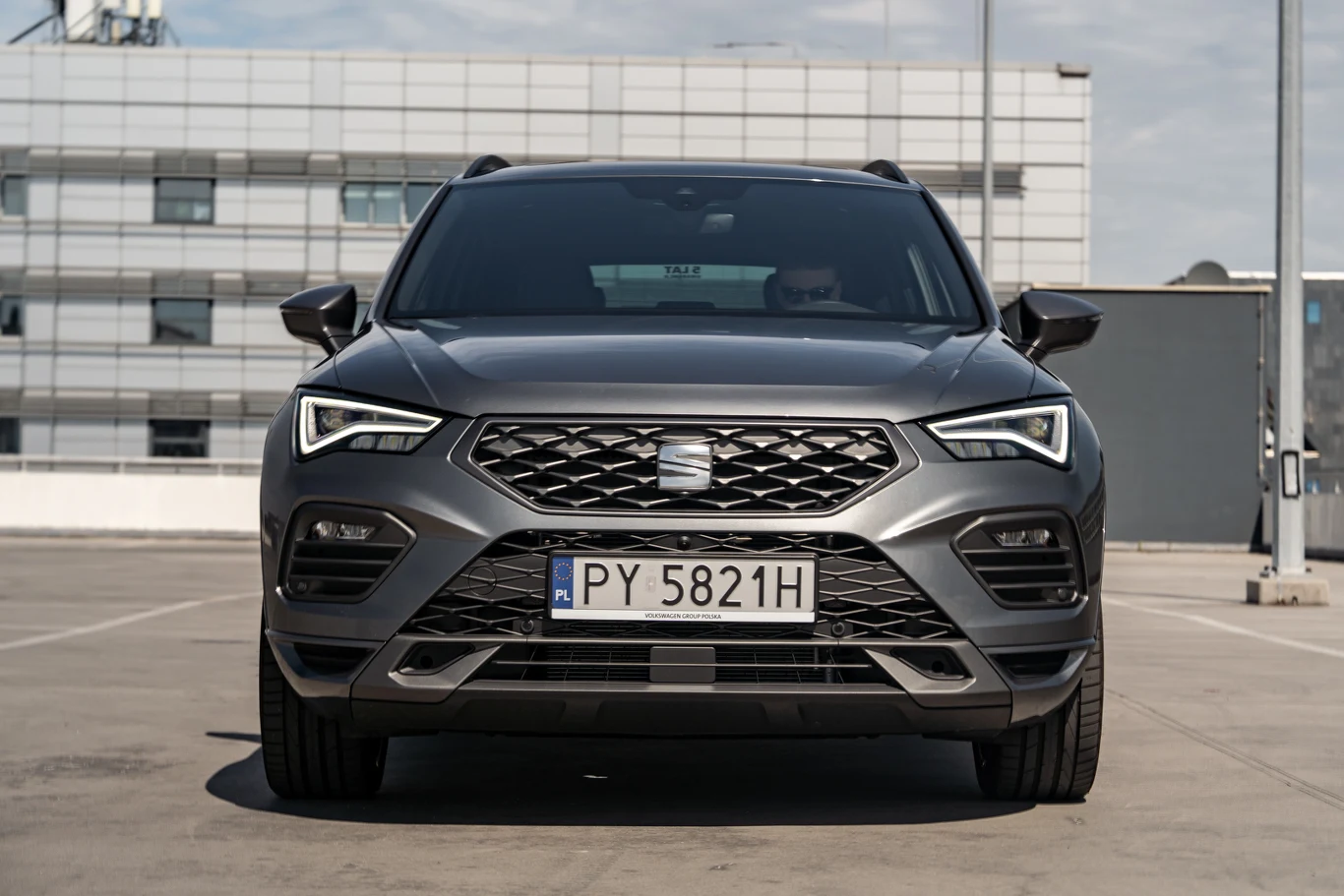 Nowoczesny samochód osobowy marki Seat szarego koloru zaparkowany na betonowym parkingu, widoczny od przodu z czytelną tablicą rejestracyjną i charakterystycznym logo na grillu, w tle budynek biurowy.