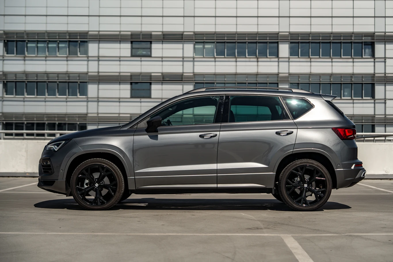 Nowoczesny, srebrny SUV z czarnymi felgami zaparkowany na wielopoziomowym parkingu, w tle widać fasadę budynku z dużymi oknami.