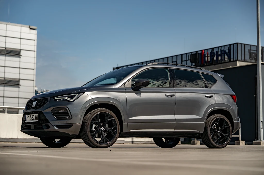 Srebrny crossover typu SUV marki Seat stoi na parkingu na tle nowoczesnego budynku z przeszkleniami i niebieskim niebem. Samochód zwraca uwagę sportową stylistyką oraz czarnymi felgami.