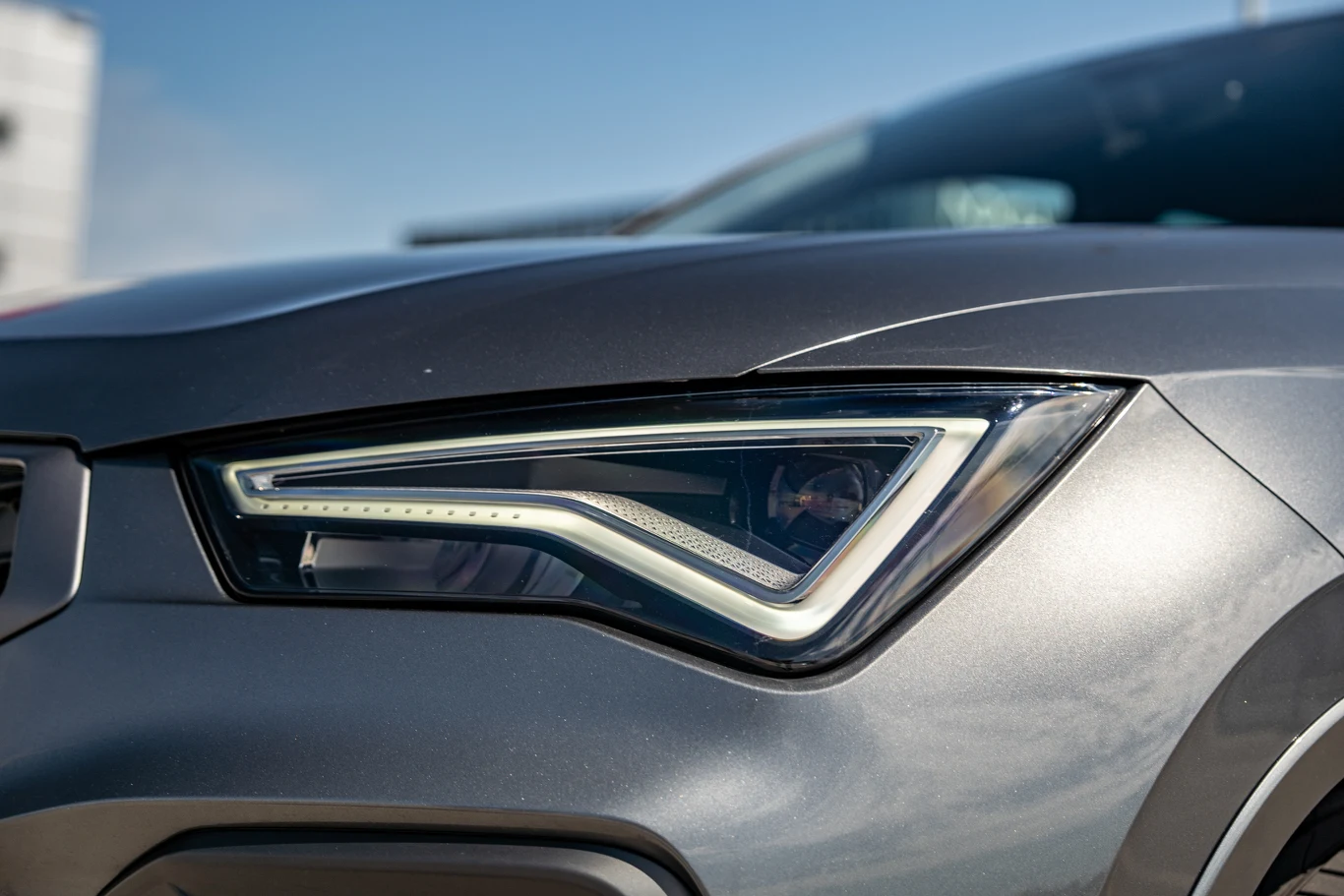 Zbliżenie na nowoczesny reflektor samochodu w szarej karoserii, z wyraźnie widocznymi detalami oświetlenia LED oraz fragmentem maski i błotnika.