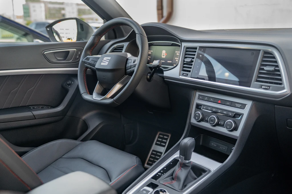 Kokpit nowoczesnego samochodu marki Seat z widoczną kierownicą, deską rozdzielczą, ekranem multimedialnym oraz panelami klimatyzacji i nawiewów. Tapicerka wykonana ze skóry, wnętrze utrzymane w eleganckim, czarnym kolorze.