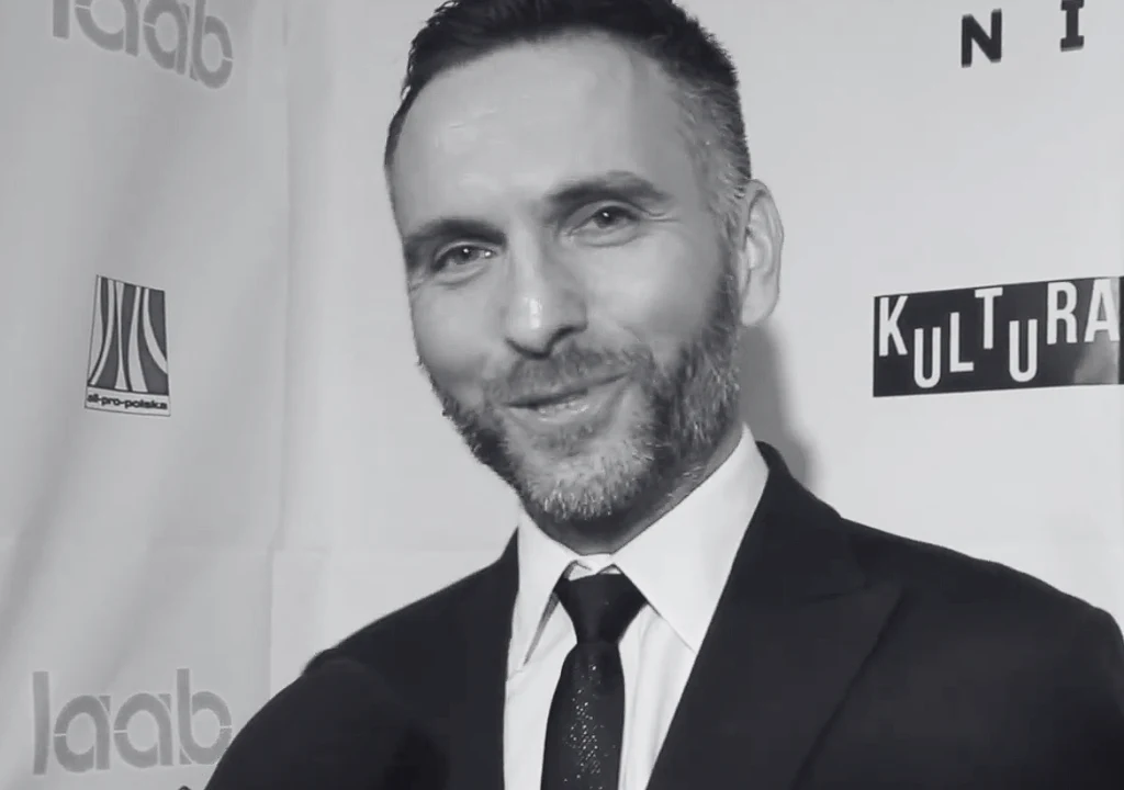 Paweł Hartlieb Uśmiechnięty mężczyzna w garniturze i krawacie stoi na tle ściany z logotypami różnych wydarzeń lub sponsorów.