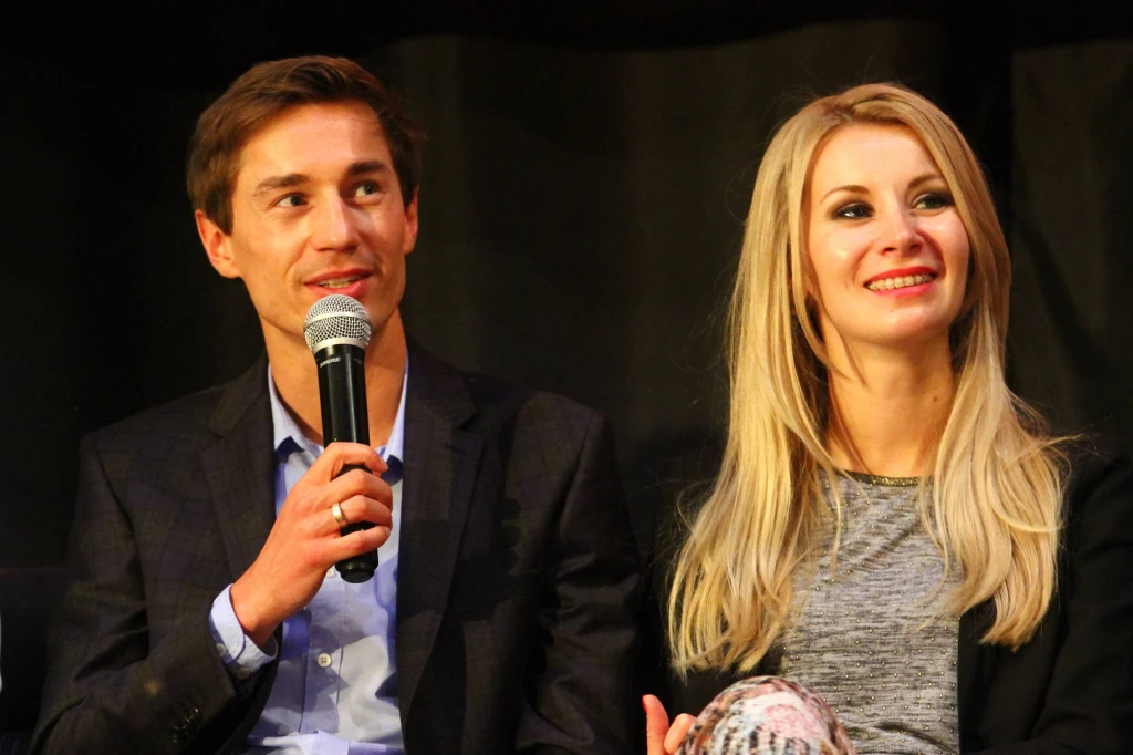 Kamil Stoch i jego małżonka Ewa Bilan-Stoch Mężczyzna w garniturze trzyma mikrofon i uśmiecha się, siedząc obok kobiety o blond włosach, która także się uśmiecha. Oboje wyglądają na przygotowanych do rozmowy lub wystąpienia publicznego.