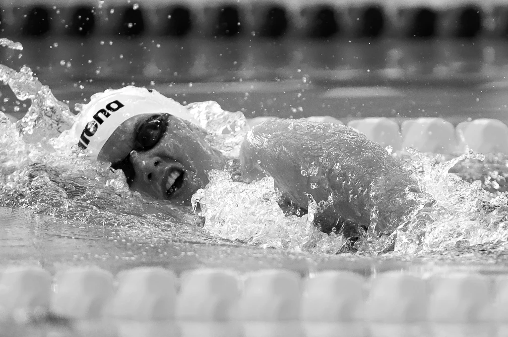 Justyna Burska Zawodnik pływacki w czepku i okularach płynie stylem dowolnym w basenie, otoczony wodnymi rozbryzgami, skoncentrowany na wysiłku sportowym.