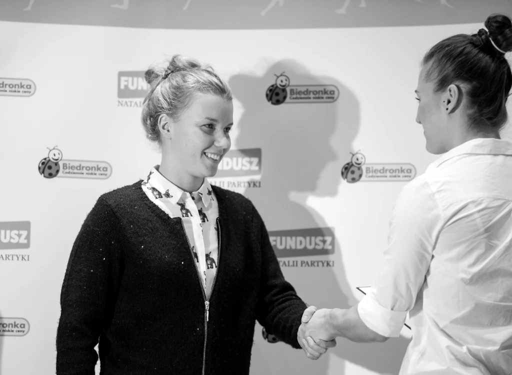 Justyna Burska Dwie kobiety podają sobie ręce w geście powitania lub gratulacji na tle ścianki z logo Fundusz Natalii Partyki oraz sieci sklepów Biedronka.