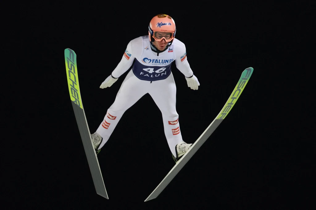 Zawodnik w białym kombinezonie narciarskim w locie podczas skoku na nartach, widoczny numer startowy 46 oraz sprzęt sportowy, tło czarne podkreślające dynamikę momentu.