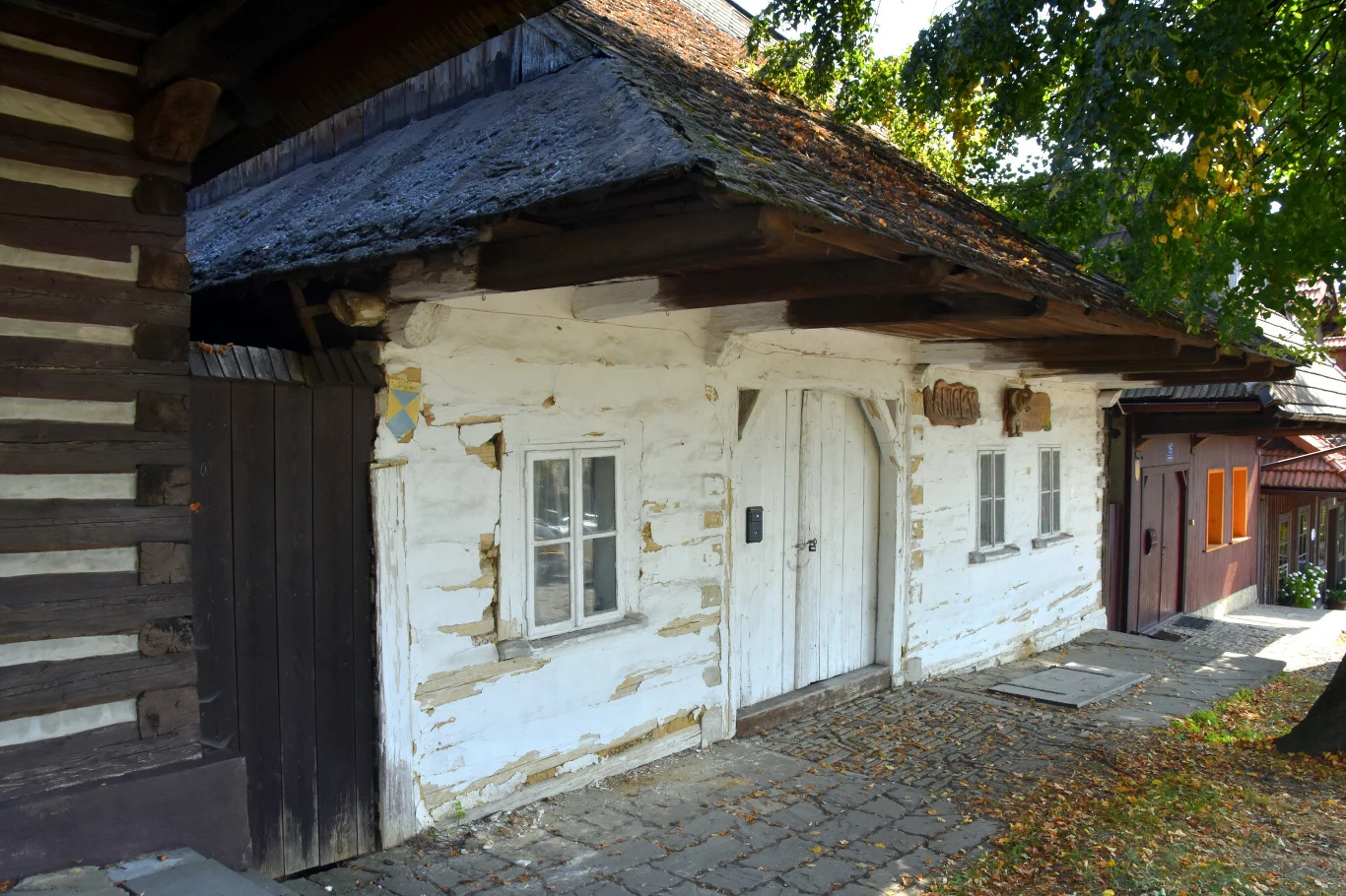Stara, drewniana chata z bielonymi ścianami i małymi oknami, dach pokryty strzechą, wokół brukowany chodnik i drzewa rzucające cień, obok widać fragment kolejnego budynku.