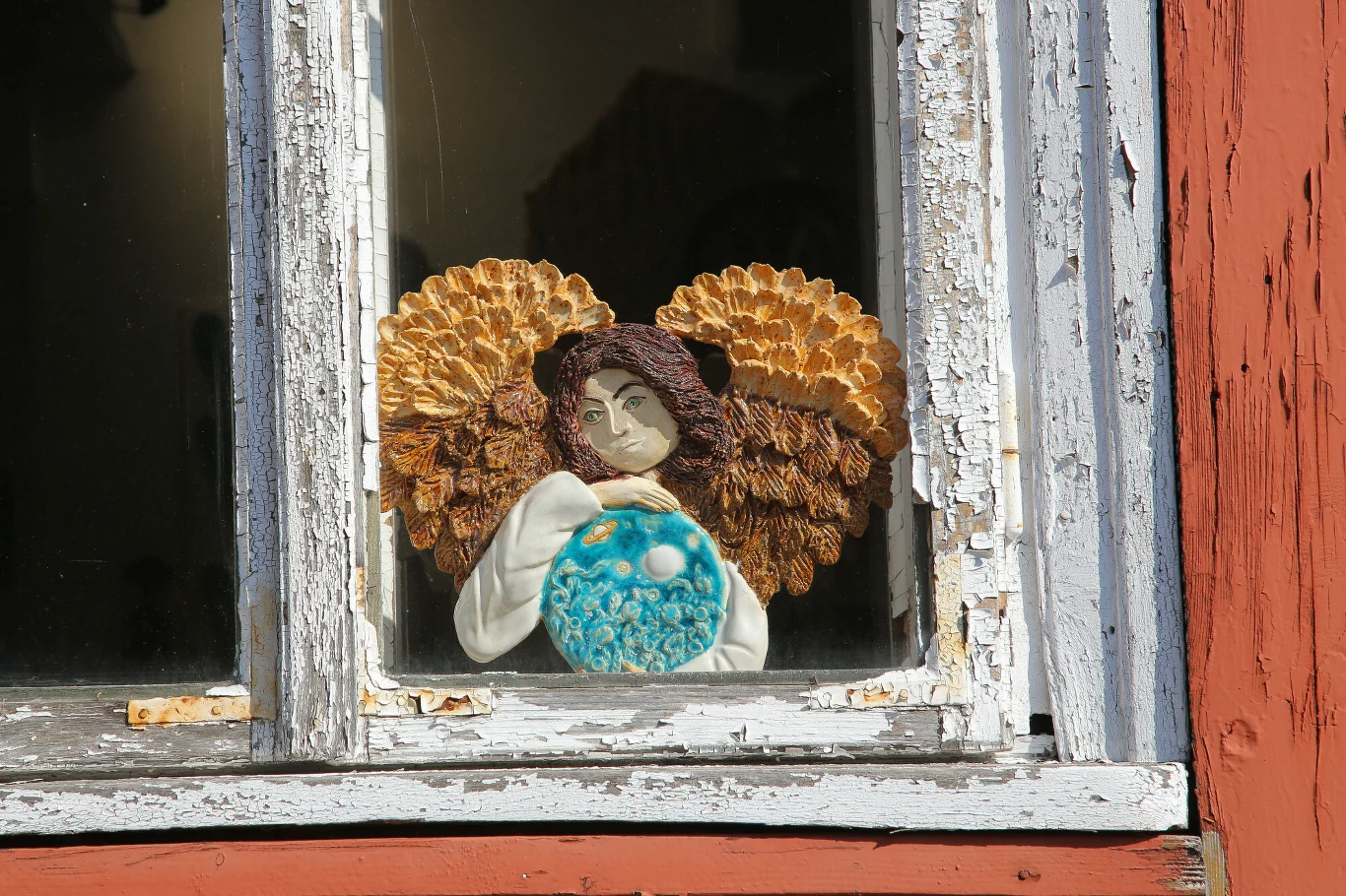 Figura anioła z dużymi złotobrązowymi skrzydłami i niebieską kulą w dłoniach ustawiona za szybą starego, częściowo obdrapanego okna. Tło stanowi ściana pomalowana na ciepły czerwony kolor i wyblakła biała rama okna.