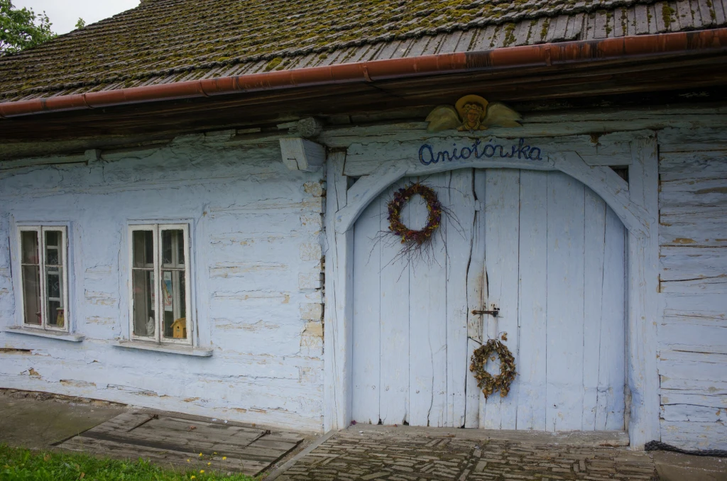 Lanckorona Drewniany, jasnobłękitny budynek z dwoma oknami po lewej stronie i dużymi, podwójnymi drzwiami ozdobionymi wieńcami, nad wejściem napis Aniołowka oraz ozdoba anioła.