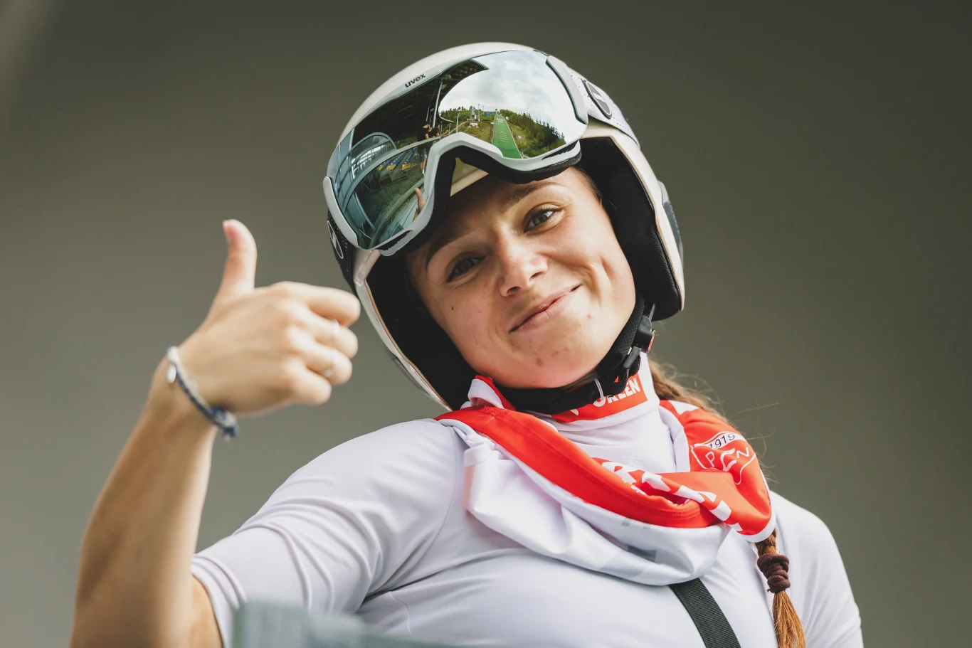 Uśmiechnięta kobieta w kasku i okularach sportowych unosi kciuk do góry, wokół szyi ma przewieszoną czerwoną chustę, w tle rozmyte tło.