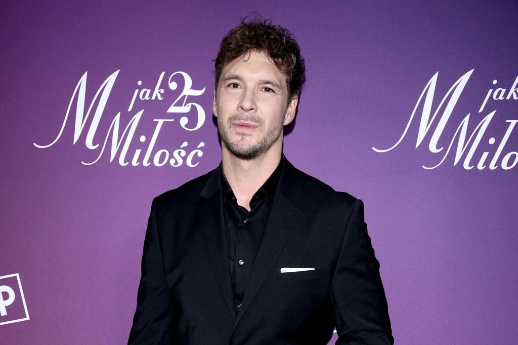 Mikołaj Roznerski na 25-leciu "M jak miłość" Mężczyzna w eleganckim, czarnym garniturze i koszuli stoi na tle fioletowej ścianki promującej jubileusz serialu "M jak Miłość". Twarz dobrze widoczna, jednolite tło z białym napisem.