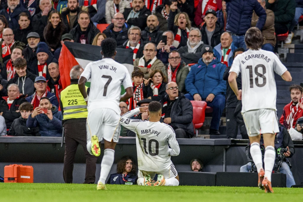 Kylian Mbappe celebruje zdobycie bramki przed sektorem zajmowanym przez kibiców gospodarzy Piłkarz w białym stroju klęczy na murawie po zdobyciu bramki, dwaj inni zawodnicy biegną w jego stronę. W tle trybuny pełne kibiców, niektórzy wyrażają emocje i robią zdjęcia.