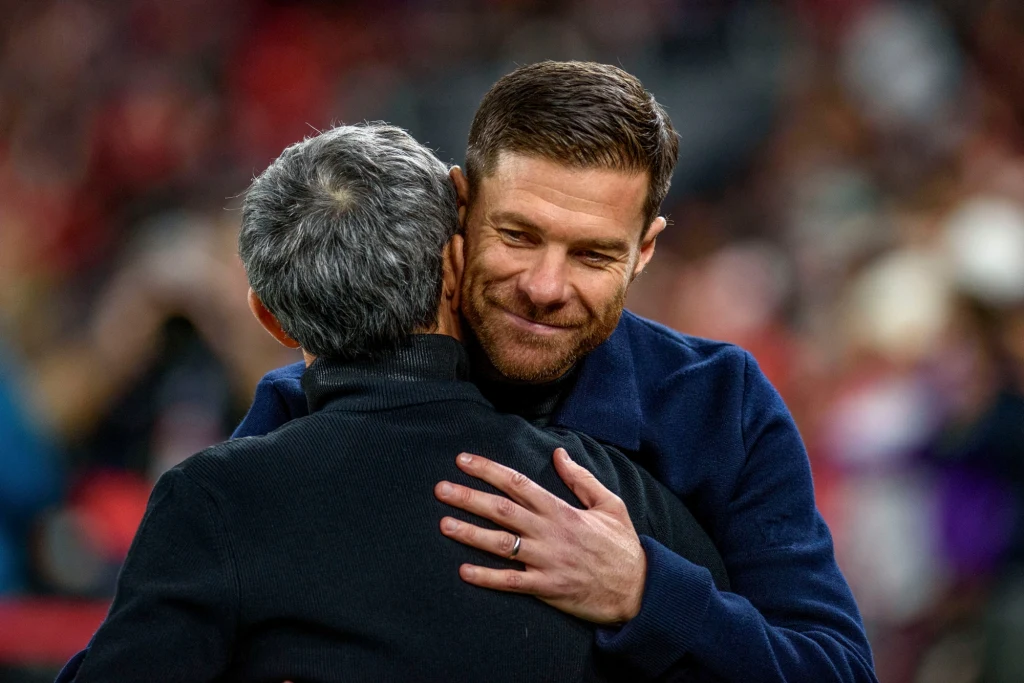 Serdeczne przywitanie Xabiego Alonso (przodem) i Ernesto Valverde Dwie osoby obejmują się w serdecznym uścisku. Jeden z mężczyzn ma lekko pochyloną głowę i przyjazny uśmiech, drugi widoczny jest od tyłu. Tło jest nieostre, co podkreśla emocjonalny charakter spotkania.