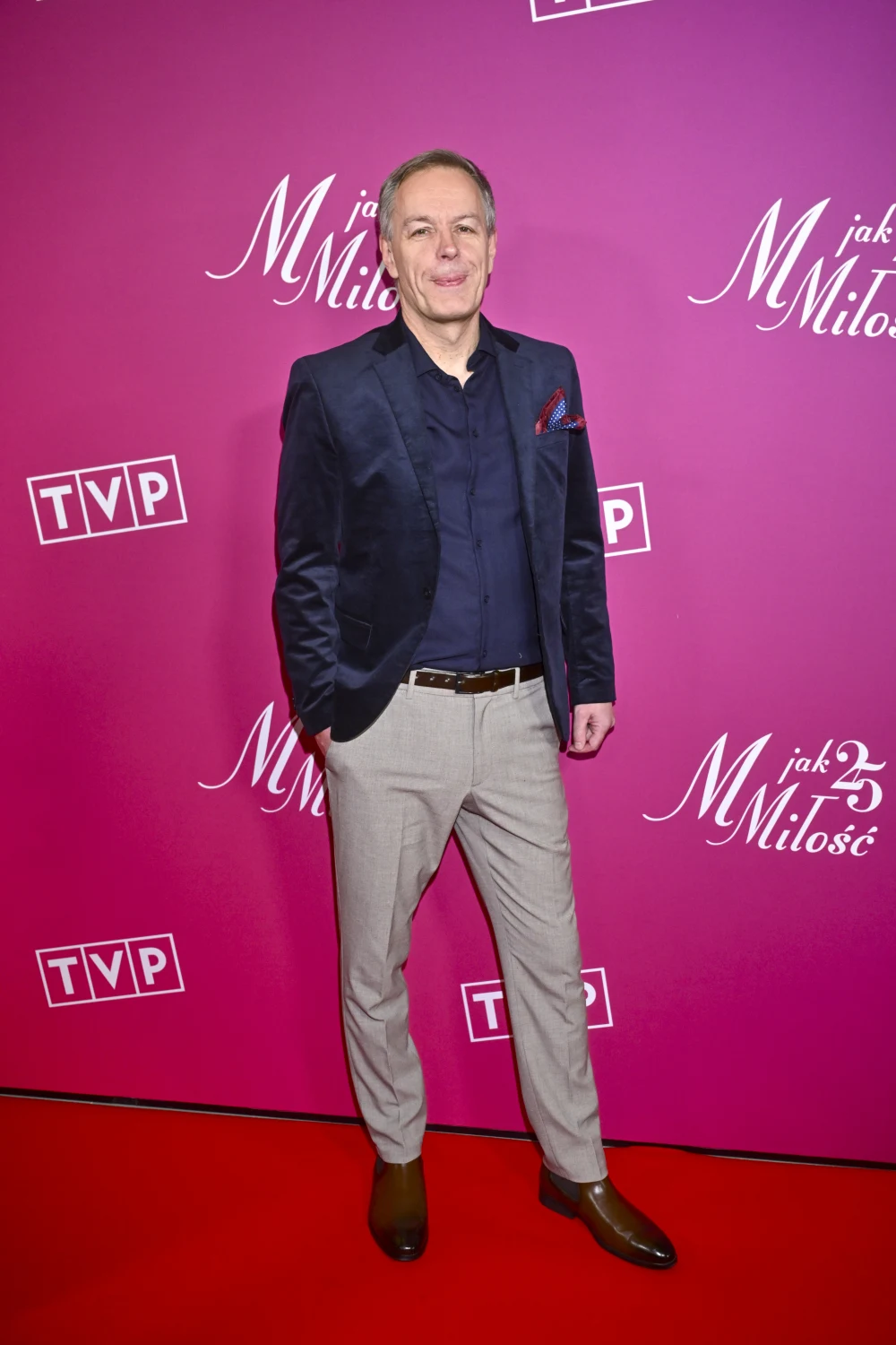 Steffen Möller na 25-leciu "M jak miłość" Ubrany elegancko mężczyzna pozuje na czerwonym dywanie, w tle fioletowa ścianka z logotypami TVP oraz 'M jak Miłość'.