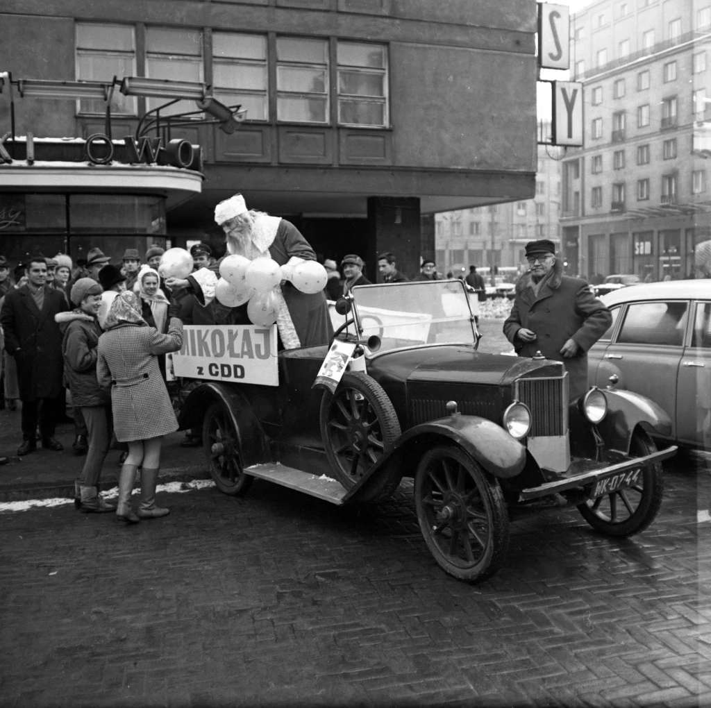 Stary samochód udekorowany balonami i tabliczką z napisem 'Mikołaj z CDD', na platformie siedzi przebrany za Mikołaja mężczyzna rozdający prezenty dzieciom, wokół grupka ludzi oraz przechodniów na miejskiej ulicy.