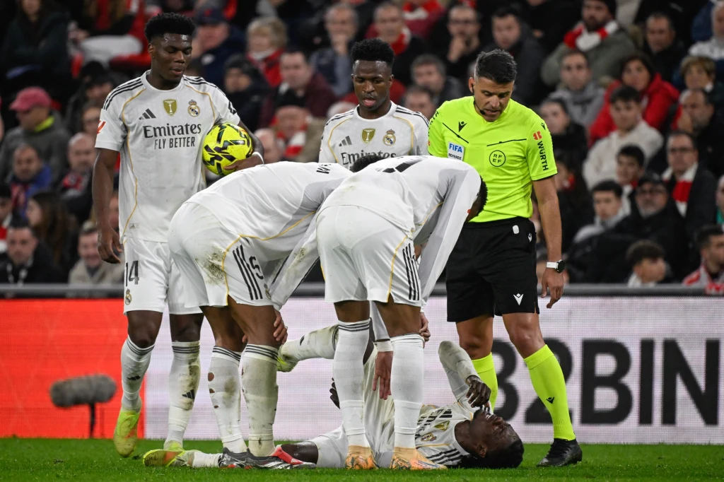 "Los Blancos" okupili triumf kontuzją Eduardo Camavingi Piłkarz leży na murawie otoczony przez swoich kolegów z drużyny w białych strojach Realu Madryt oraz sędziego ubranych w żółty strój, podczas meczu rozgrywanego przy pełnych trybunach.