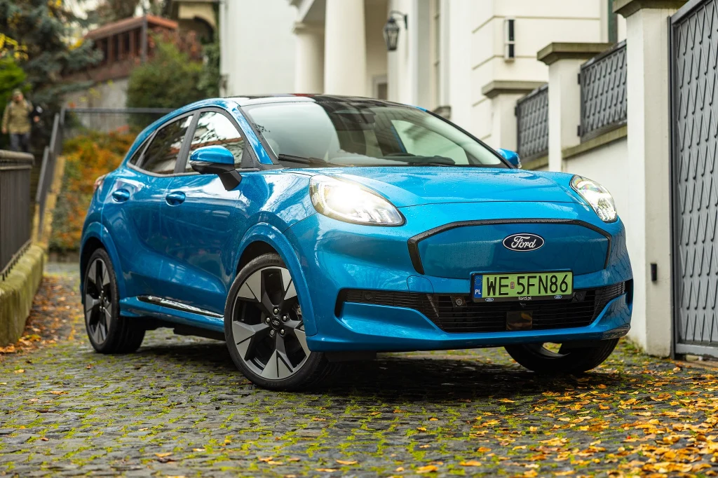Ford Puma Gen-E to najmniejszy elektryczny model Forda Jasnoniebieski samochód marki Ford stoi zaparkowany na brukowanej ścieżce otoczonej liśćmi, przed wejściem do stylowego budynku; w tle widoczne drzewa oraz fragmenty sąsiedniej zabudowy.