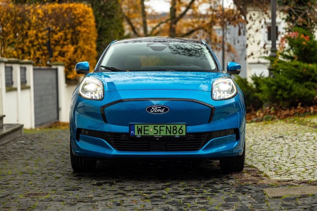 Gdyby nie zielona tablica rejestracyjna, elektryczną Pumę można by pomylić ze spalinową wersją Jasnoniebieski samochód osobowy marki Ford na brukowanej nawierzchni, widoczny od przodu, otoczenie stanowi ozdobny ogród z jesienną roślinnością i elementami architektury ogrodowej.