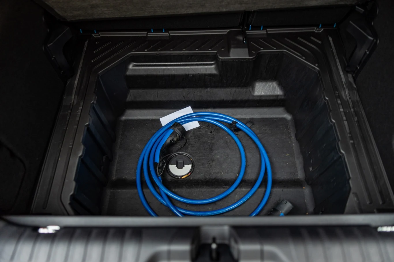 Czarny schowek w bagażniku samochodu z niebieskim przewodem do ładowania pojazdu elektrycznego, ułożonym spiralnie, w centralnej części przestrzeni.
