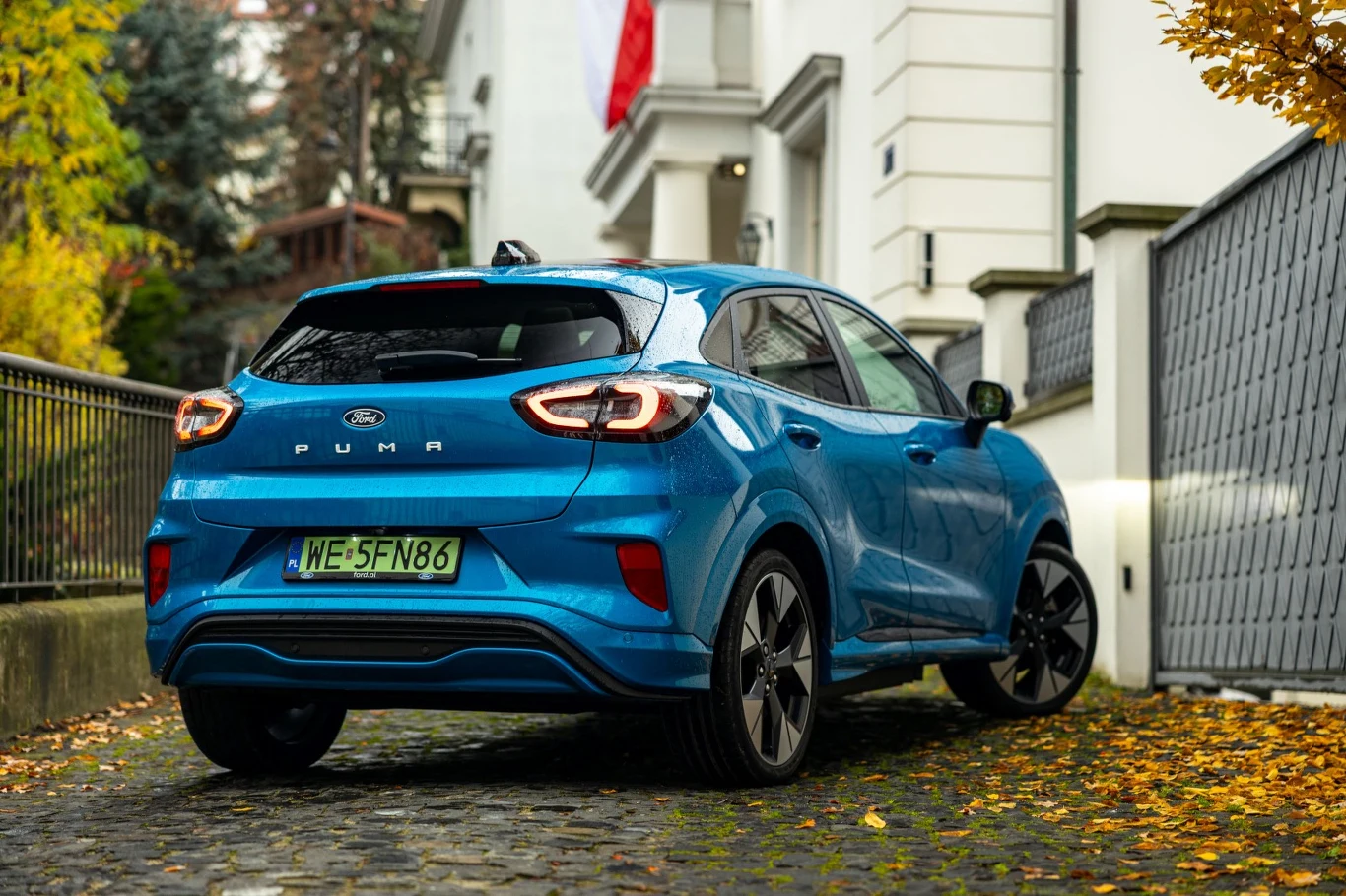 Niebieski samochód Ford Puma zaparkowany na brukowanej alejce w otoczeniu jesiennych liści, przy białym budynku z polską flagą na fasadzie.