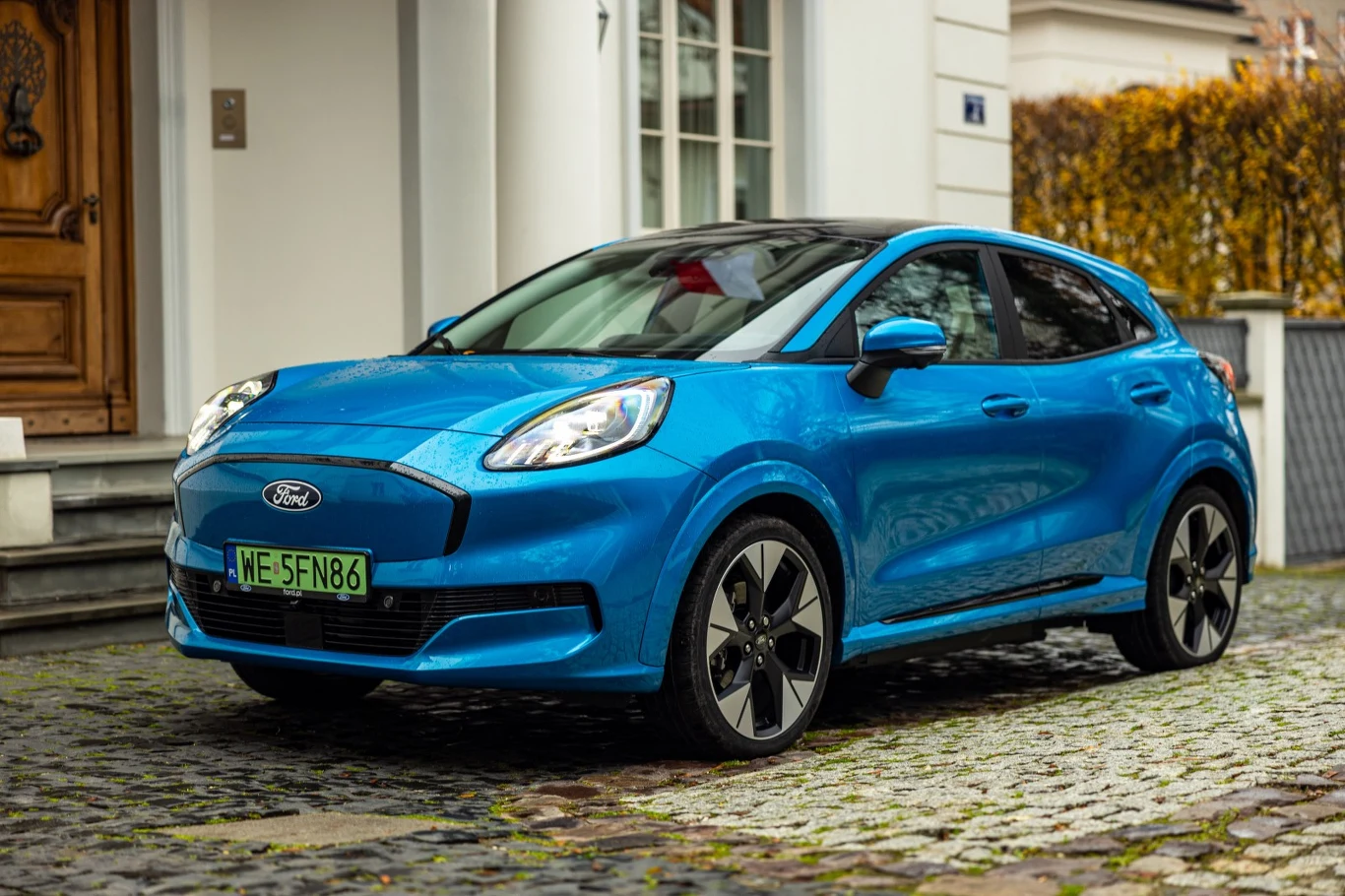 Niebieski samochód Ford Puma zaparkowany na brukowanej ulicy przed wejściem do eleganckiego budynku mieszkalnego.