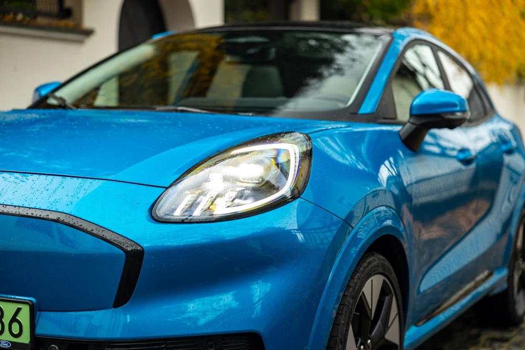 Puma Gen-E w wersji Premium została wyposażona w reflektory matrycowe Nowoczesny, niebieski samochód osobowy zaparkowany przy ulicy, widoczny fragment przedniej części pojazdu z podświetlonym reflektorem, na karoserii krople deszczu.