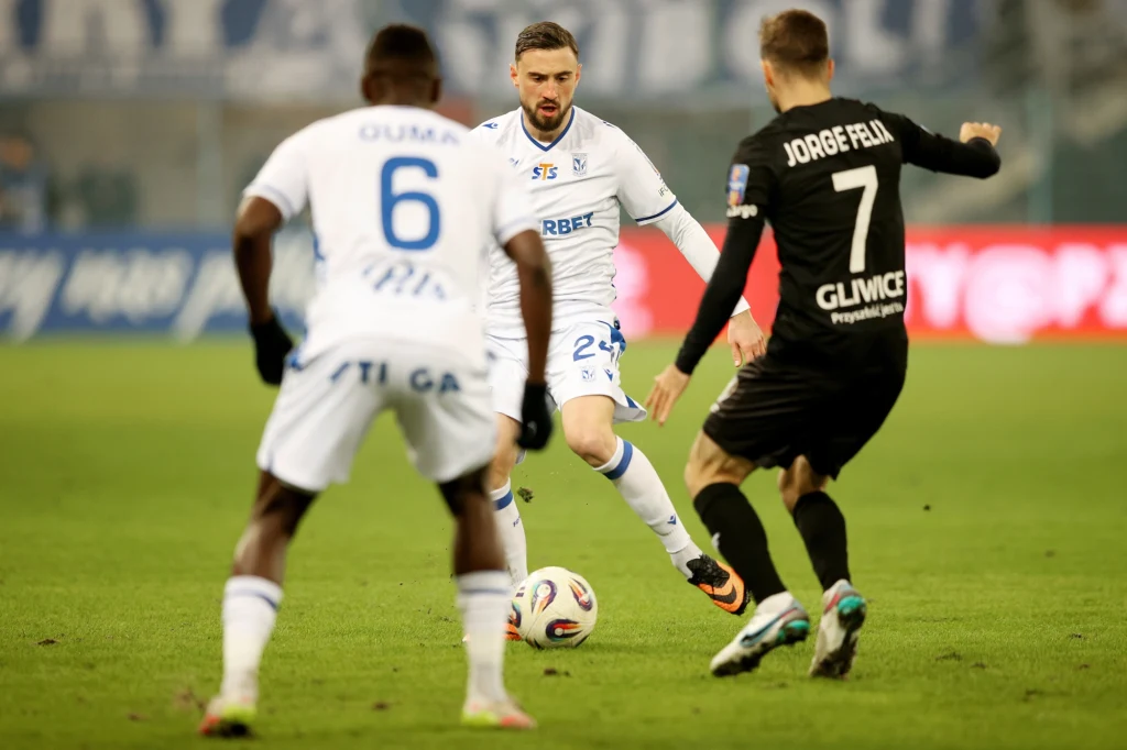 Piast Gliwice - Lech Poznań w Pucharze Polski Zawodnik w białej koszulce drybluje piłką, naprzeciwko niego stoją rywale, w tym jeden w czarnej koszulce z napisem Jorge Felix. Piłkarze skupieni na akcji na boisku podczas meczu piłki nożnej.