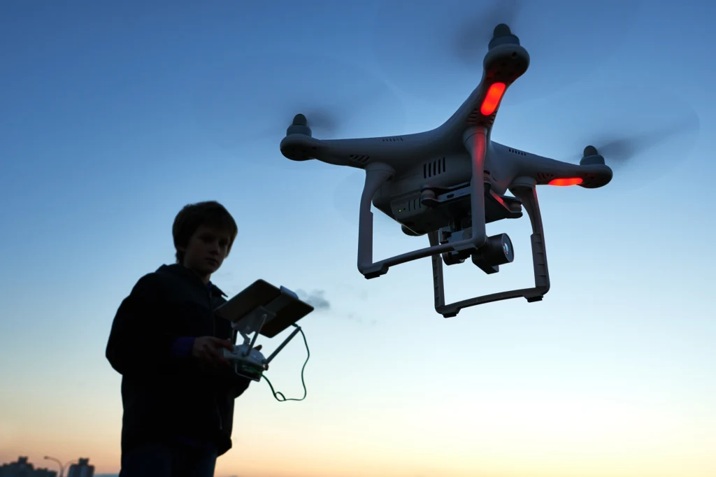 Droniada Junior pokazuje, że drony to nauka przyszłości Chłopiec sterujący dronem przy zachodzącym słońcu, duży dron z kamerą w powietrzu, wyraźnie widoczne światła sygnalizacyjne na ramionach urządzenia.