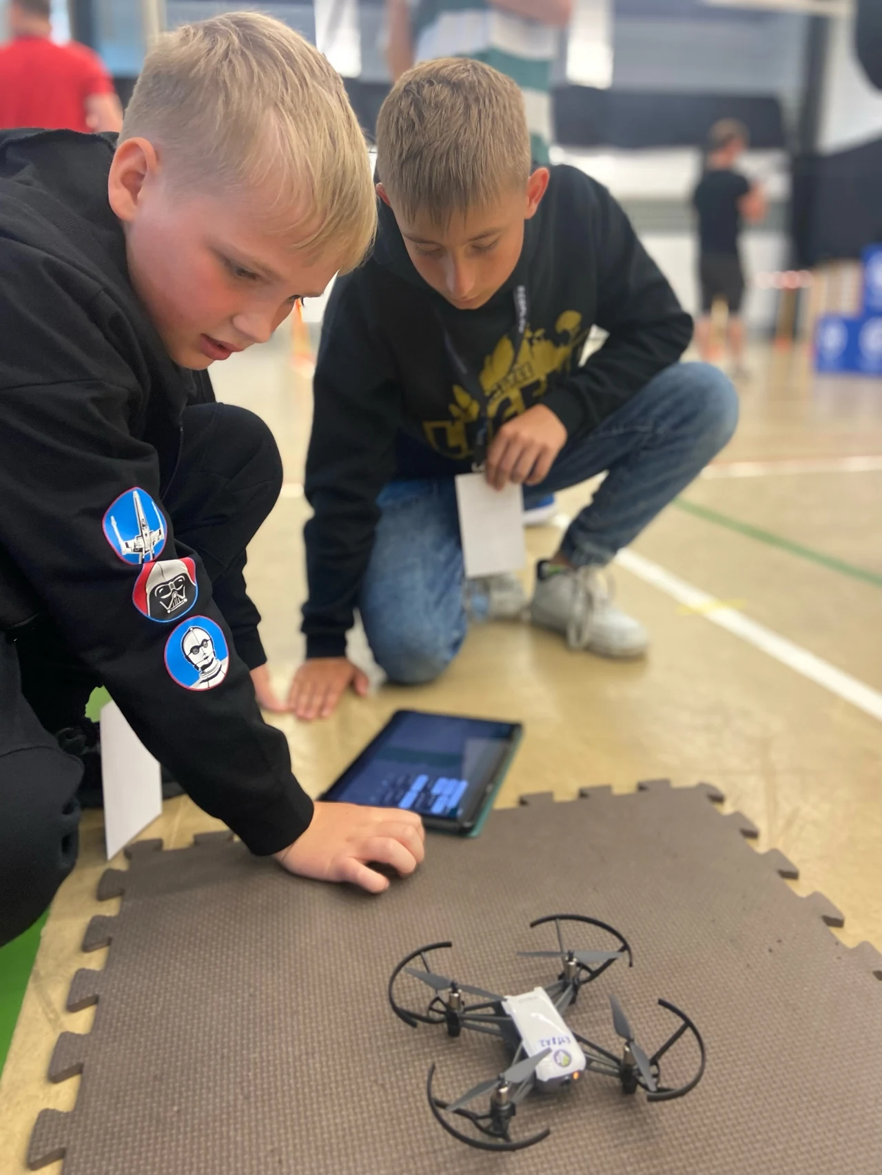 Szkoły odkrywają drony. Droniada Junior może odmienić lekcje Dwóch chłopców koncentruje się na małym dronie leżącym na macie, obok tabletu z aplikacją sterującą. Jeden z nich wskazuje na drona, drugi kontroluje ekran tabletu.