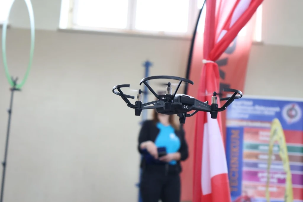 Turniej nie jest przeznaczony tylko dla szkół z doświadczeniem w dronach. Dron unoszący się w powietrzu w pomieszczeniu, w tle rozmyta postać trzymająca tablet lub kontroler oraz elementy dekoracyjne takie jak flaga, plansze informacyjne i obręcz.