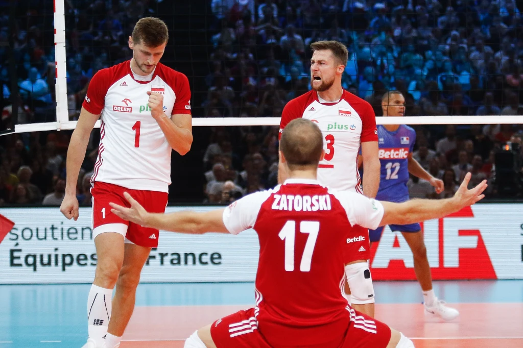 Polscy siatkarze w biało-czerwonych strojach celebrują udaną akcję podczas meczu, z zawodnikiem po francuskiej stronie siatki widocznym w tle. Trybuny wypełnione kibicami podkreślają atmosferę sportowego wydarzenia.