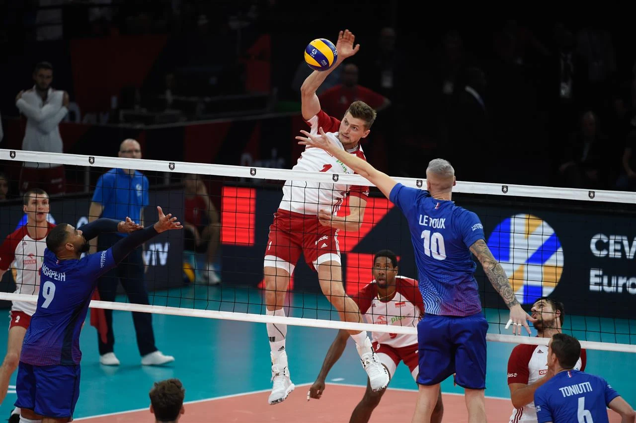 Dynamiczna akcja meczu siatkówki, zawodnik w biało-czerwonym stroju wyskakuje do ataku nad siatką, próbując zdobyć punkt, podczas gdy gracze w niebieskich strojach próbują zablokować piłkę.