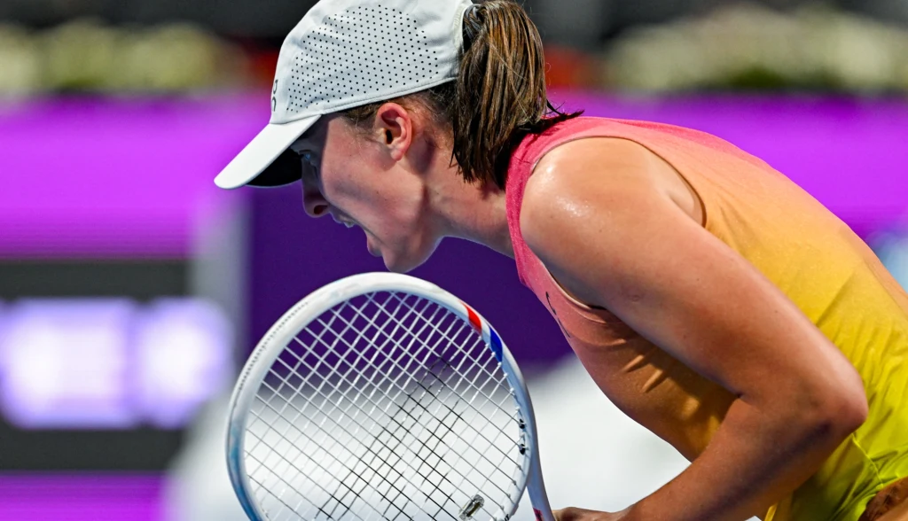 Iga Świątek Zawodniczka tenisowa w sportowym stroju i czapce, pochylona z rakietą tenisową w dłoni, skoncentrowana podczas intensywnego momentu gry, w tle rozmyty fioletowy baner.