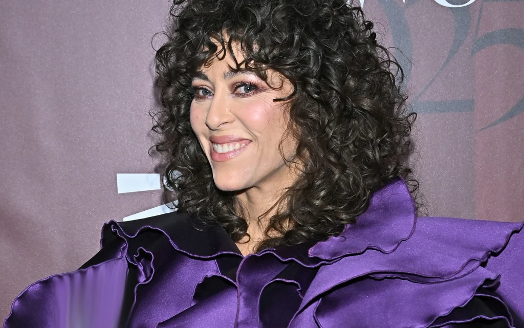 Natalia Kukulska Uśmiechnięta kobieta o ciemnych, kręconych włosach ubrana w fioletową, ozdobną suknię na tle bordowej ściany z elementami graficznymi.