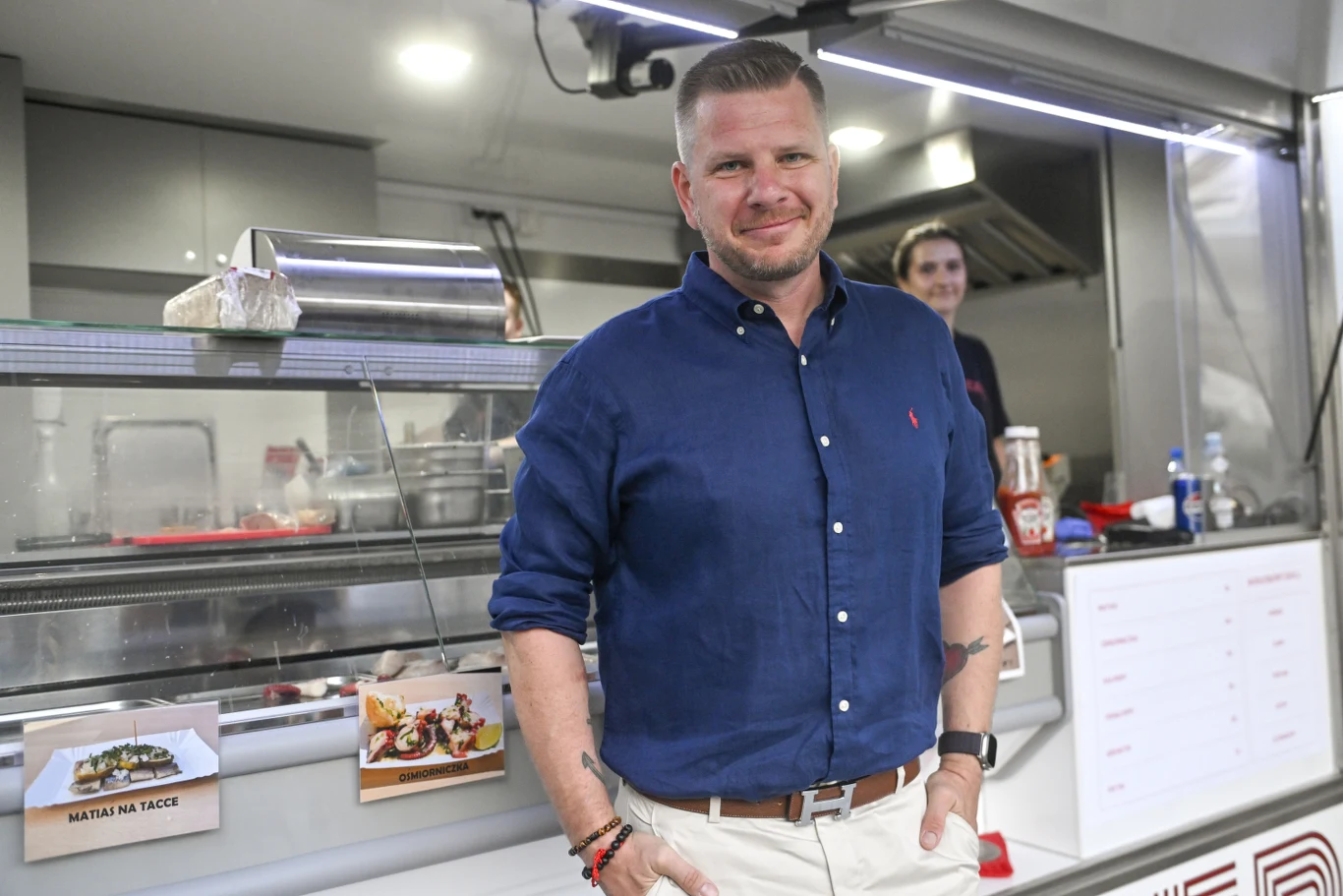 Mężczyzna w granatowej koszuli i jasnych spodniach stoi uśmiechnięty przed food truckiem, w tle widoczna jest druga osoba oraz elementy wyposażenia kuchni, na ladzie znajdują się przykładowe potrawy na zdjęciach.