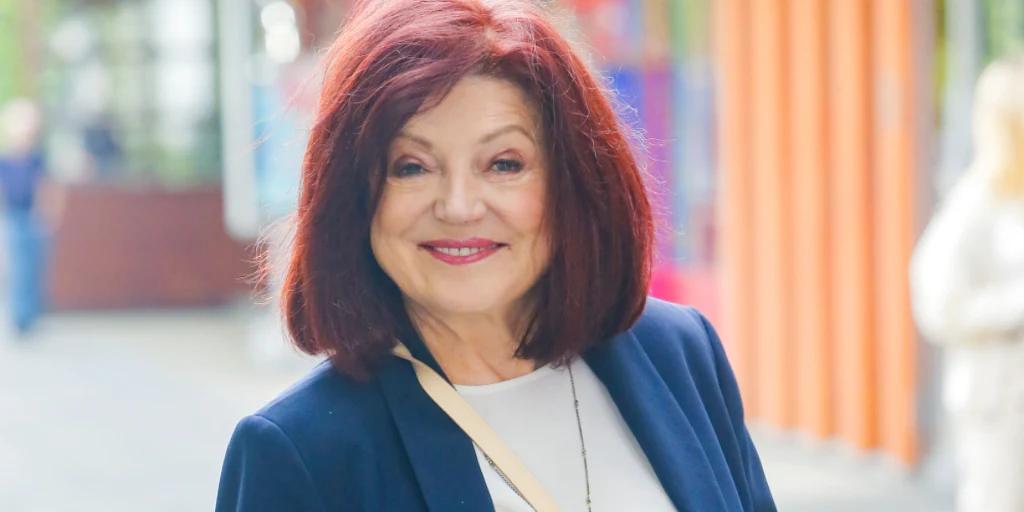 Urszula Dudziak Kobieta w średnim wieku z rudymi włosami uśmiecha się, stojąc na tle kolorowych murów na zewnątrz. Ubrana w niebieską marynarkę i jasną bluzkę z przewieszoną torebką przez ramię.