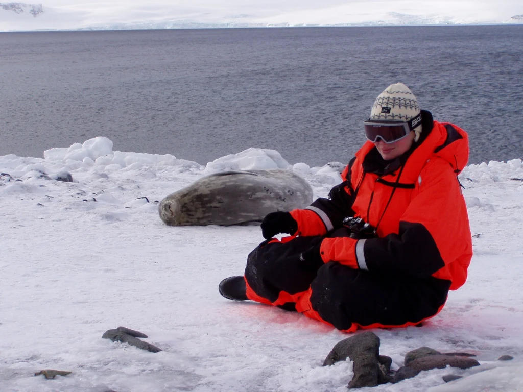Osoba w jasnopomarańczowym kombinezonie zimowym siedzi na śniegu nad brzegiem zamarzniętego akwenu, w pobliżu leży foka odpoczywająca na lodzie.