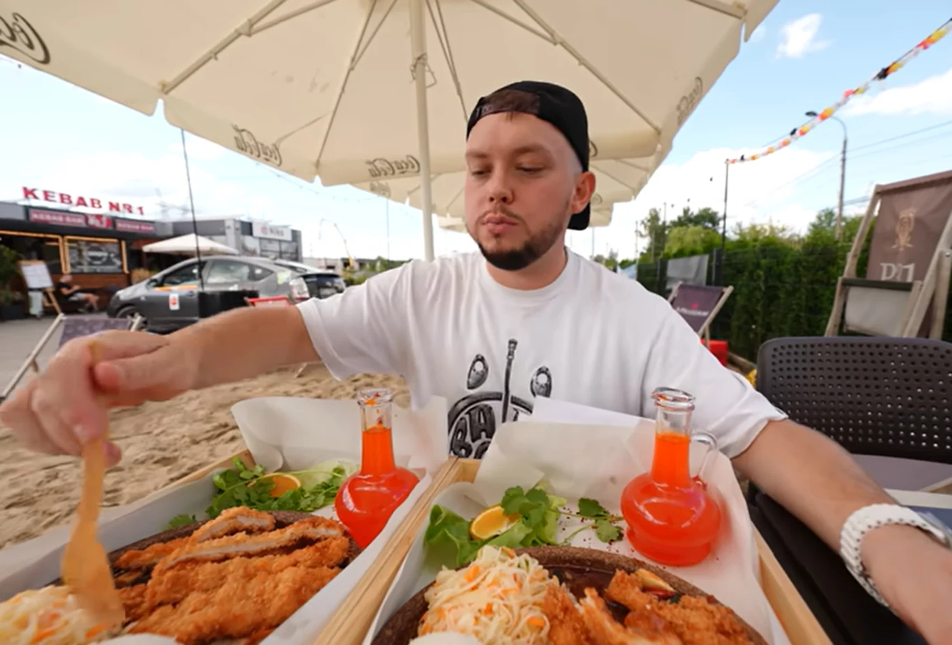 Mężczyzna w białej koszulce siedzi przy stole na zewnątrz, pod parasolem, i sięga po jedzenie. Na stole dwa talerze z panierowanym kurczakiem, surówką oraz dwie butelki z pomarańczowym napojem. W tle widoczne są napisy reklamowe oraz samochody.