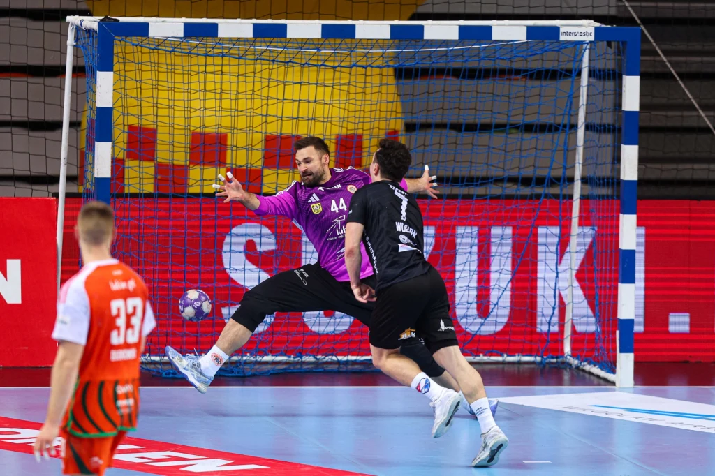 Dynamiczna akcja pod bramką podczas meczu piłki ręcznej, bramkarz w fioletowym stroju próbuje obronić rzut, a inny zawodnik w czarnym stroju znajduje się tuż przed nim, w tle widoczni kolejni gracze i bramka.